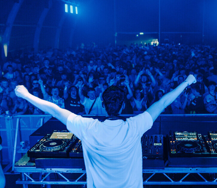 Chris Flannigan performs on the Pumphouse stage at AVA Belfast 2025, raising his arms to a full crowd illuminated in blue light.