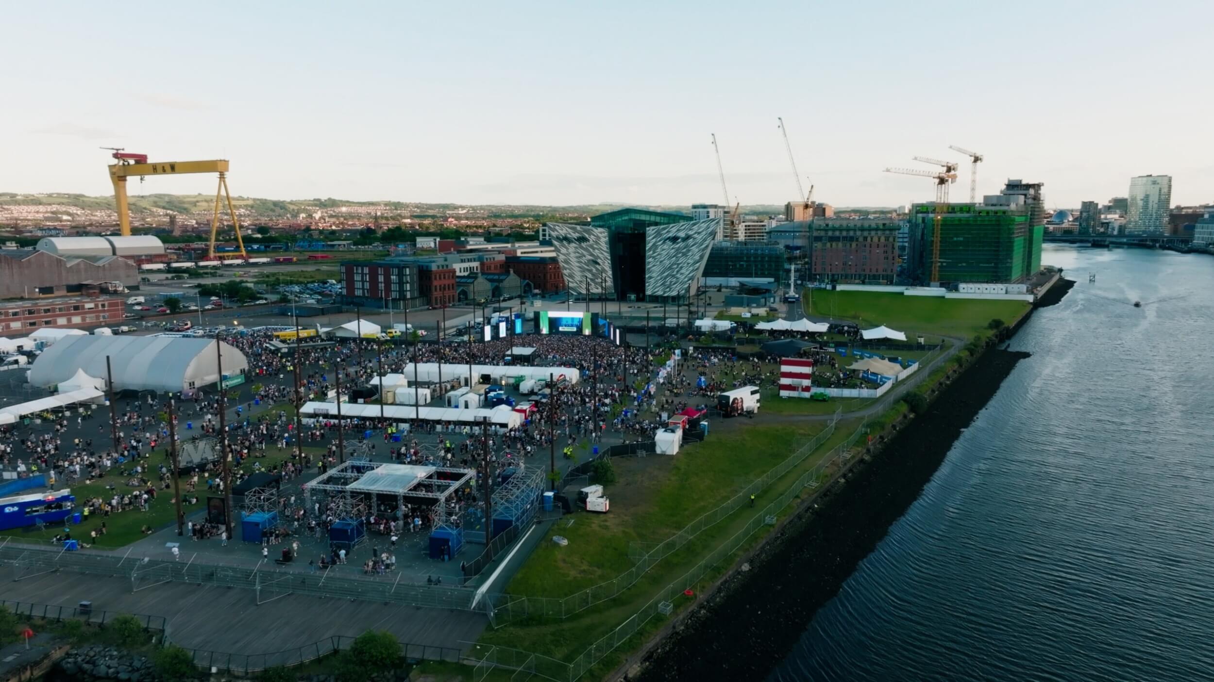 Drone aerial view of AVA Belfast Festival 2025, showing large crowd, stage lighting, and festival structures in an open outdoor venue in Belfast.